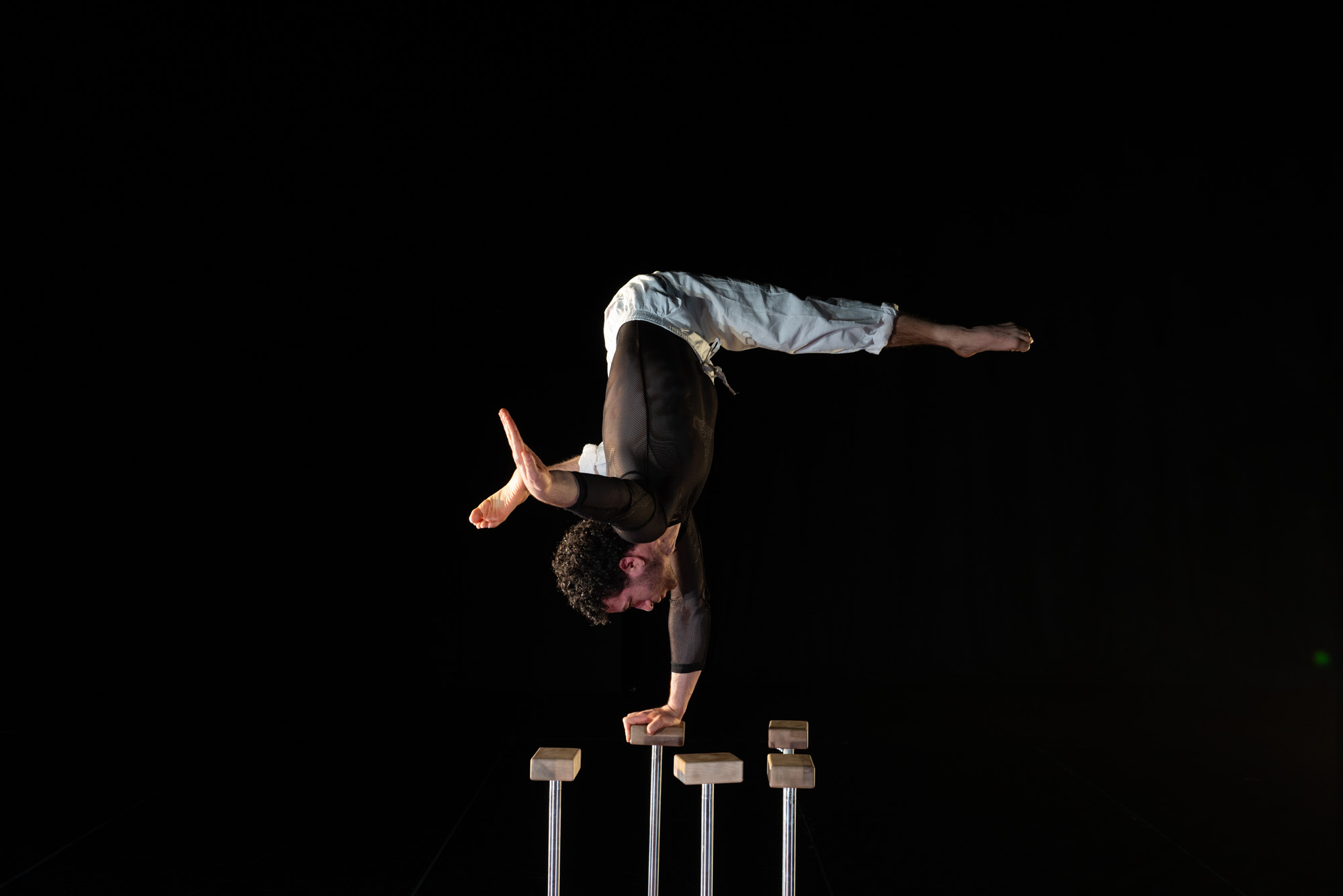 Un acrobate masculin est en équilibre sur ses mains au sommet de quatre barres parallèles, les jambes écartées en grand écart, sur un fond sombre.