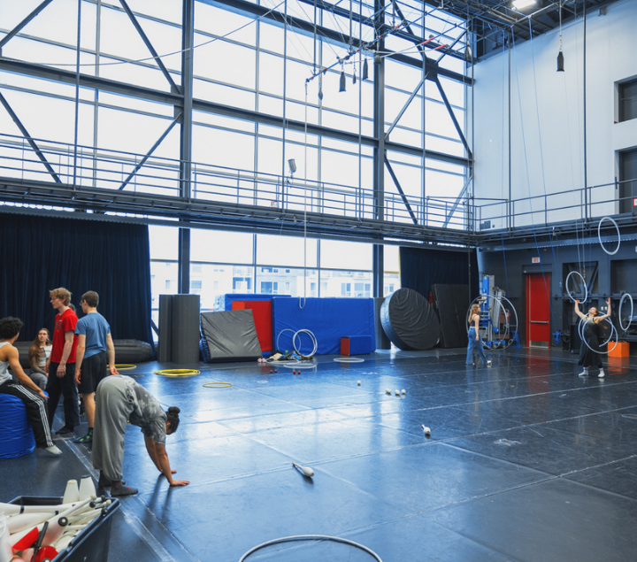 Les participants s'entraînent aux disciplines circassiennes dans un gymnase spacieux doté de hauts plafonds, de grandes baies vitrées, de cerceaux aériens et de tapis disséminés un peu partout. Certains jonglent, d'autres font des étirements ou des exercices d'équilibre, dans un espace d'entraînement coloré et bien éclairé.
