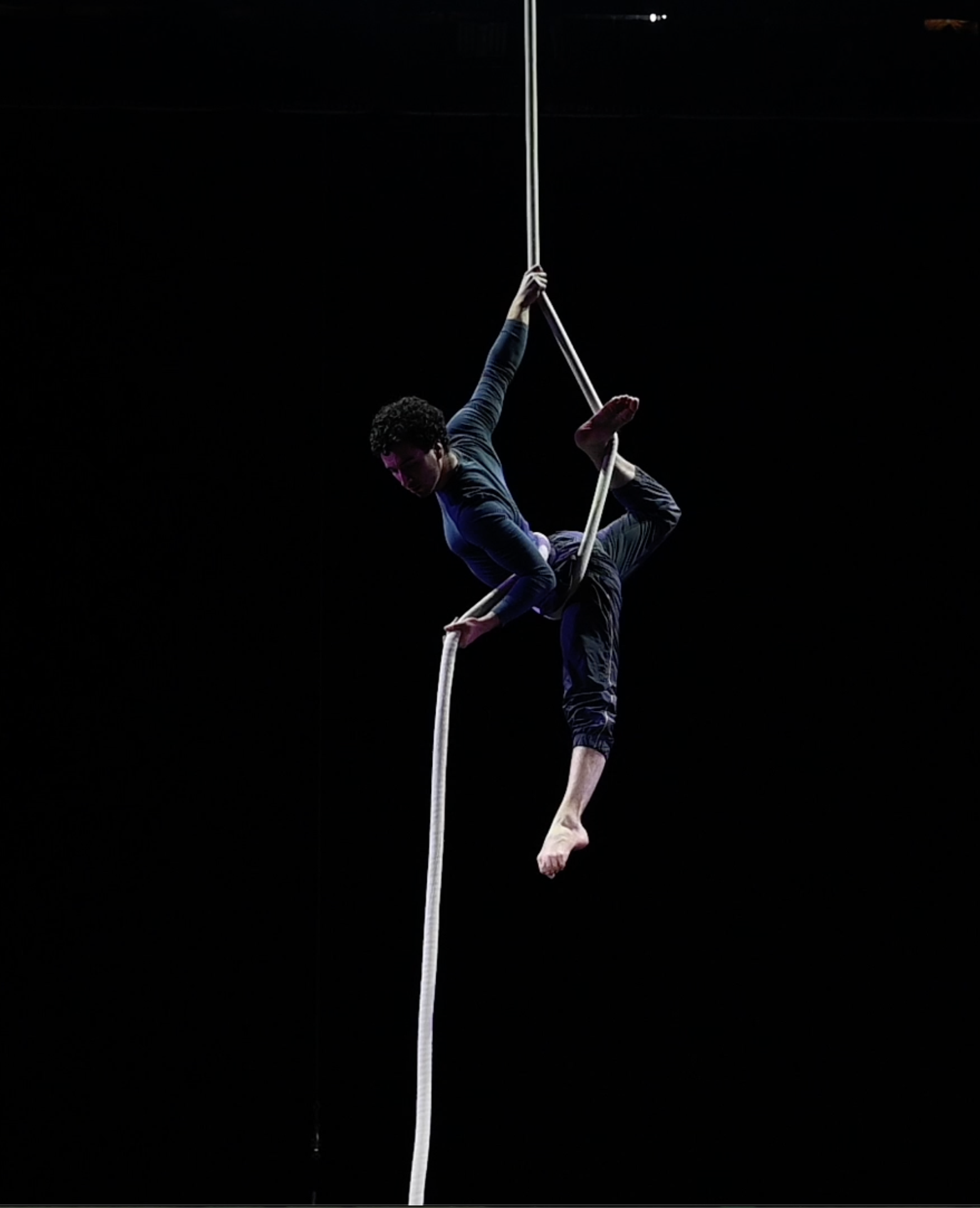 Une personne exécute un numéro d'acrobatie aérienne, suspendue et en équilibre sur une corde blanche, sur fond sombre.