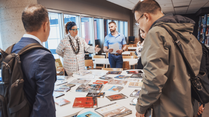 Dans une bibliothèque, un groupe de personnes est rassemblé autour d'une table recouverte de livres et de magazines. L'une d'entre elles gesticule tout en parlant, tandis que les autres l'écoutent attentivement ; on aperçoit des étagères et des fenêtres en arrière-plan.