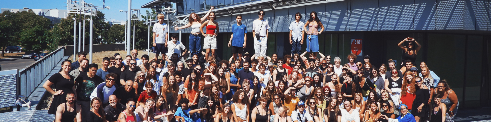 Un groupe nombreux et hétéroclite de personnes pose ensemble devant un bâtiment moderne par une journée ensoleillée ; certaines se tiennent debout sur une structure située derrière la foule principale, tandis que d’autres font des gestes ludiques ou sourient à l’appareil photo, illustrant ainsi l’esprit dynamique de ceux qui se lancent dans une carrière dans le milieu des arts du cirque.