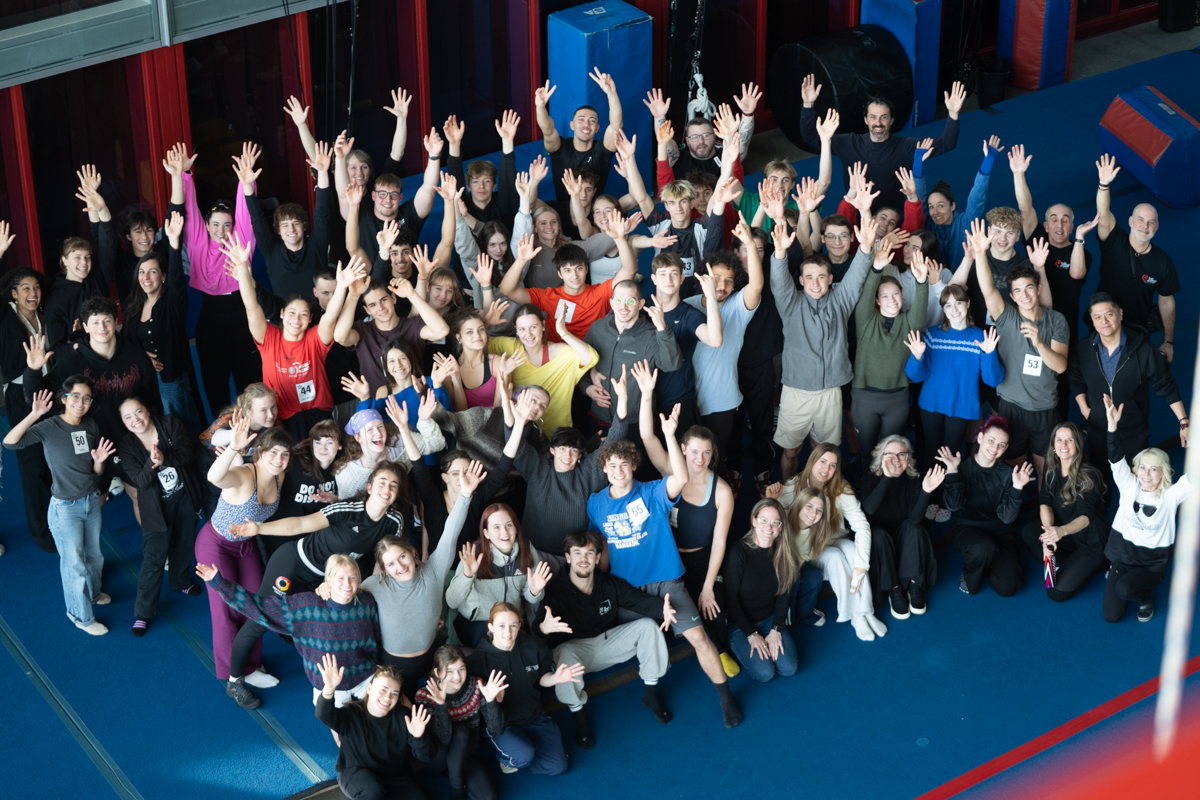 Un grand groupe de personnes pose ensemble à l'intérieur, sur un sol bleu, souriant et saluant la caméra avec enthousiasme lors des auditions de l'ÉNC.