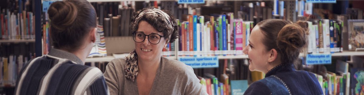Trois femmes sont assises autour d'une table ronde dans une bibliothèque, en pleine conversation. On aperçoit en arrière-plan des étagères remplies de livres colorés. Elles semblent détendues et s'échangent des sourires ; elles discutent peut-être des initiatives du Bureau d'intervention et de prévention.