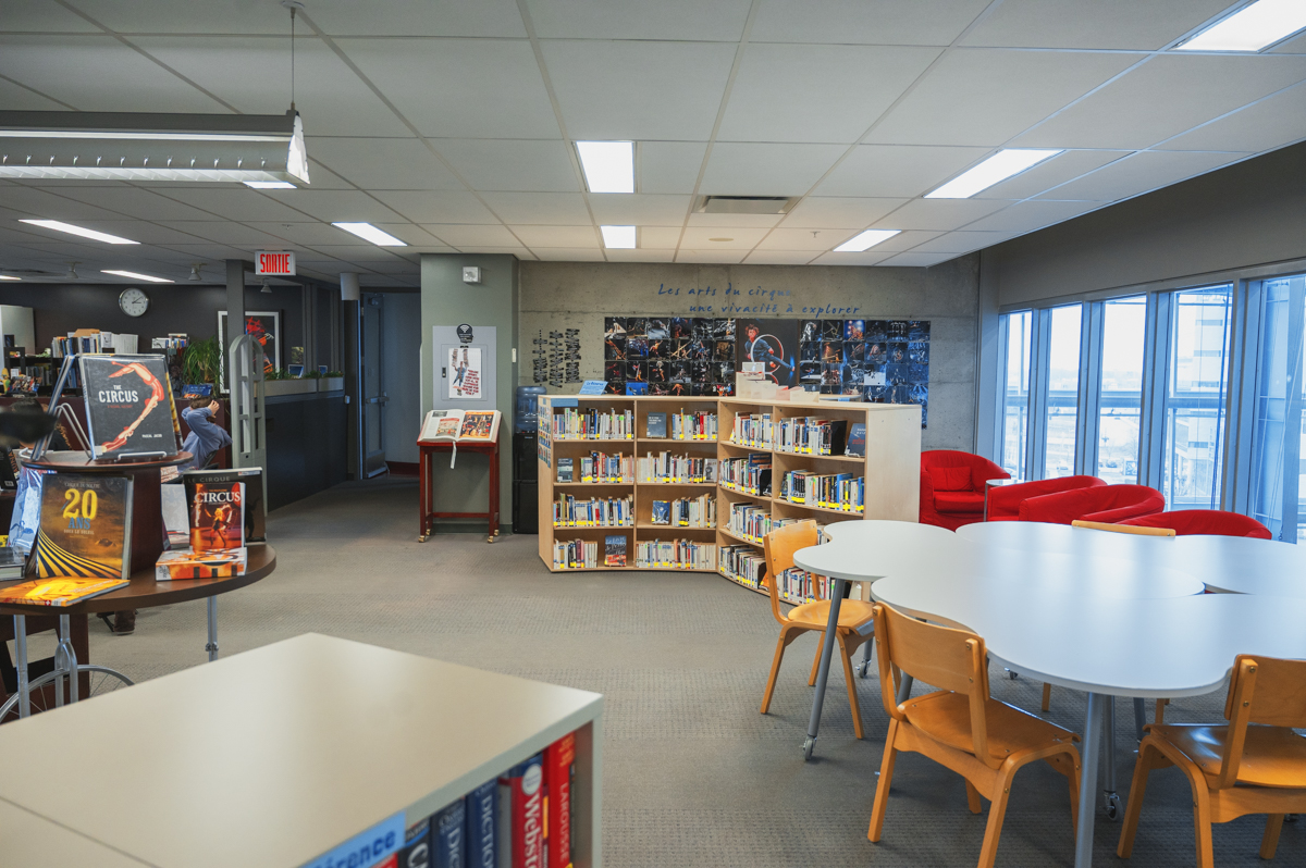 La Bibliothèque de l'ENC, un espace moderne, est agrémentée d'étagères, de fauteuils rouges, de tables rondes entourées de chaises en bois et de grandes baies vitrées laissant entrer la lumière naturelle. Un panneau mural présente des photos et une citation, créant ainsi un espace de lecture accueillant.