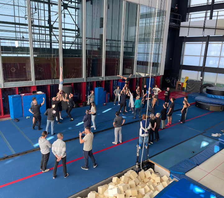 Un groupe de personnes se trouve dans un gymnase ; certaines regardent, d'autres aident une personne à être soulevée vers le plafond ; on aperçoit des tapis bleus et une fosse remplie de mousse.