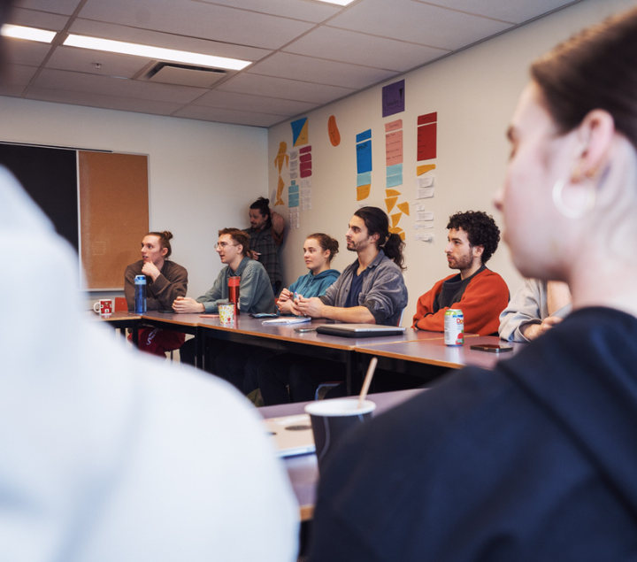 Un groupe de jeunes adultes est assis à des pupitres dans une salle de classe et écoute attentivement une personne qui ne figure pas dans le cadre. Des feuilles de couleur sont accrochées au mur derrière eux.