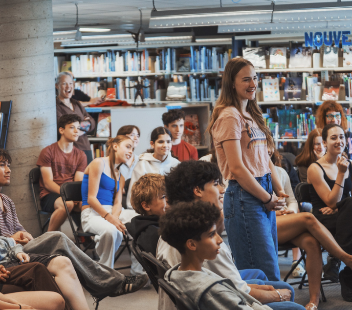 Un groupe d'adolescents et d'adultes est assis dans une bibliothèque, le sourire aux lèvres, à l'écoute d'une personne hors champ. Une jeune femme se tient debout, l'air captivée. Des étagères remplissent l'arrière-plan.